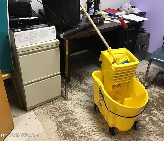Office desk, safe, and file cabinet with dry sewage on the floor and a yellow janitorial mob and bucket.
